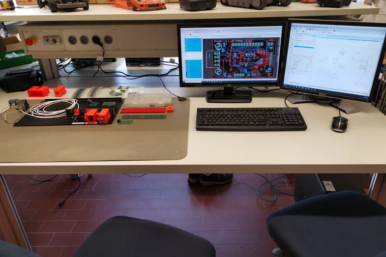 Foto von einem Arbeitsplatz mit einem Elektronikaufbau und zwei Monitoren, auf denen Arbeitsprogramme geöffnet sind.