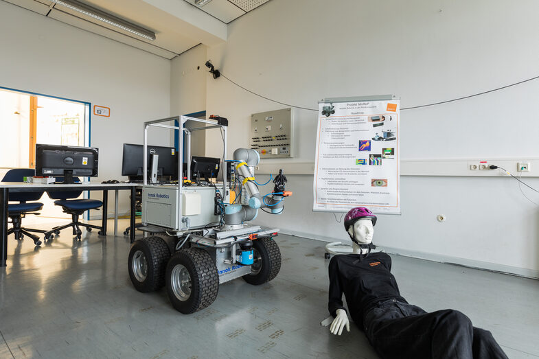 Foto von einem Rettungsroboter und Puppe mit Helm in einem Laborraum, im Hintergrund sind mehrere Monitore und oben an der Wand ist eine Kamera befestigt.