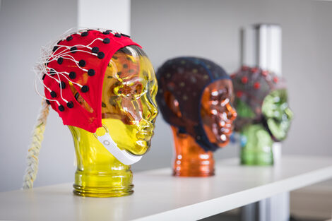 Photo of three different EEG masks with a stand in the shape of a head on a shelf. In focus is a yellow glass head on the far left. __ <br>Various EEG masks with stands in the shape of a face are lined up on a shelf. The focus is on the yellow glass head at the beginning of the row.