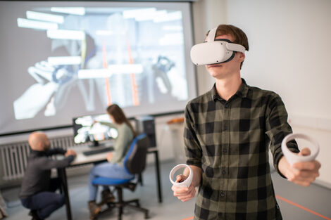 Photo of a person wearing VR goggles and holding the controllers. In the background you can see a split screen on the wall and two people talking to each other.