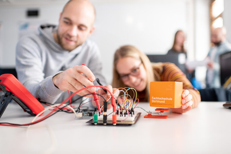 Foto von zwei Studierende, die neben einander sitzen. Der Student steckt Kabel an eine Messvorrichtung, die auf dem Tisch liegt. Diese ist an eine kleine Waage angeschlossen. Die Studentin legt den FH-Würfel darauf. __ Two students try their hand at biomedical engineering. You take a measurement with a scale. The scales are in the foreground of the picture, behind which the students sit at the table. The man holds measuring devices in his hand, his gaze wanders to the left to a measuring device. The young woman to his right is more stooped from the posture and looks at the scales, in her left hand she holds a FH cube, which she leads in the direction of the scales.