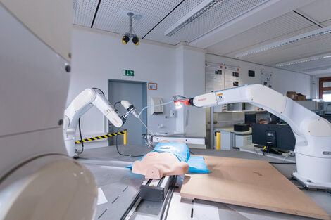 Photo of a surgical set-up with body and two surgical robots in action. The one on the right has an endoscope, the one on the left has light bulbs attached. Behind it you can see desks in the laboratory.