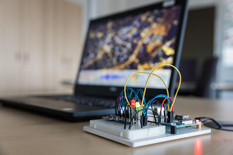 Photo of an embedded system in the foreground with activated lamps; the laptop connected to the system can be seen blurred in the background.