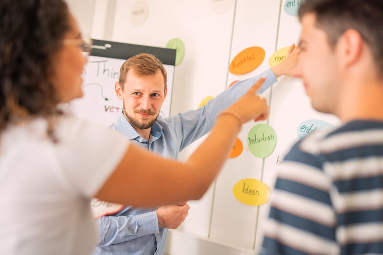 Foto von 3 Personen, die vor einer Wand stehen, an der Karten mit handgeschriebenen Worten stehen. Die Frau in der Mitte zeigt auf eine Karte.